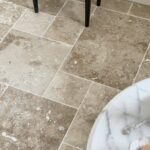 Podłoga z trawertynu CLASSIC MIX w układzie rzymskim, w ciepłych beżowo-brązowych odcieniach, z widocznymi naturalnymi porami i użyleniami. W tle jasne meble kuchenne i pleciony stołek, a na pierwszym planie marmurowy stolik z miseczkami pełnymi makaroników.