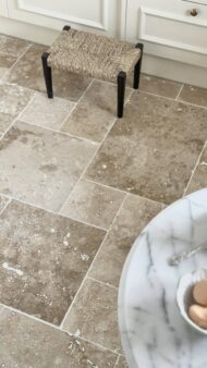 Podłoga z trawertynu CLASSIC MIX w układzie rzymskim, w ciepłych beżowo-brązowych odcieniach, z widocznymi naturalnymi porami i użyleniami. W tle jasne meble kuchenne i pleciony stołek, a na pierwszym planie marmurowy stolik z miseczkami pełnymi makaroników.