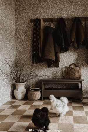 Podłoga z trawertynu PHILADELPHIA & NOUGAT w klasycznym wzorze szachownicy, wykonana z płytek 20,3x20,3 cm w beżowo-brązowych tonacjach. W tle rustykalna ściana z tapetą w liściasty wzór drewniana ławka i wieszaki na płaszcze. Na pierwszym planie jedwabiste kury silki w czarnym i białym kolorze.