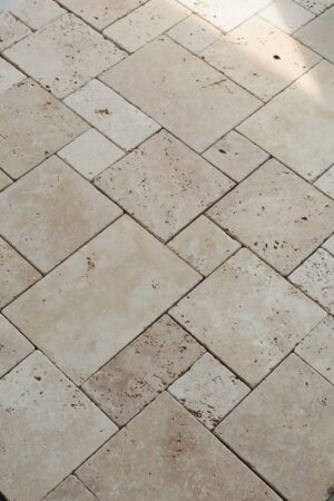 Podłoga z trawertynu IVORY MIX MINI w układzie rzymskim, złożona z płytek w ciepłych beżowych tonacjach z widocznymi porami i naturalnymi przebarwieniami. Światło podkreśla fakturę kamienia, nadając przestrzeni przytulny charakter.