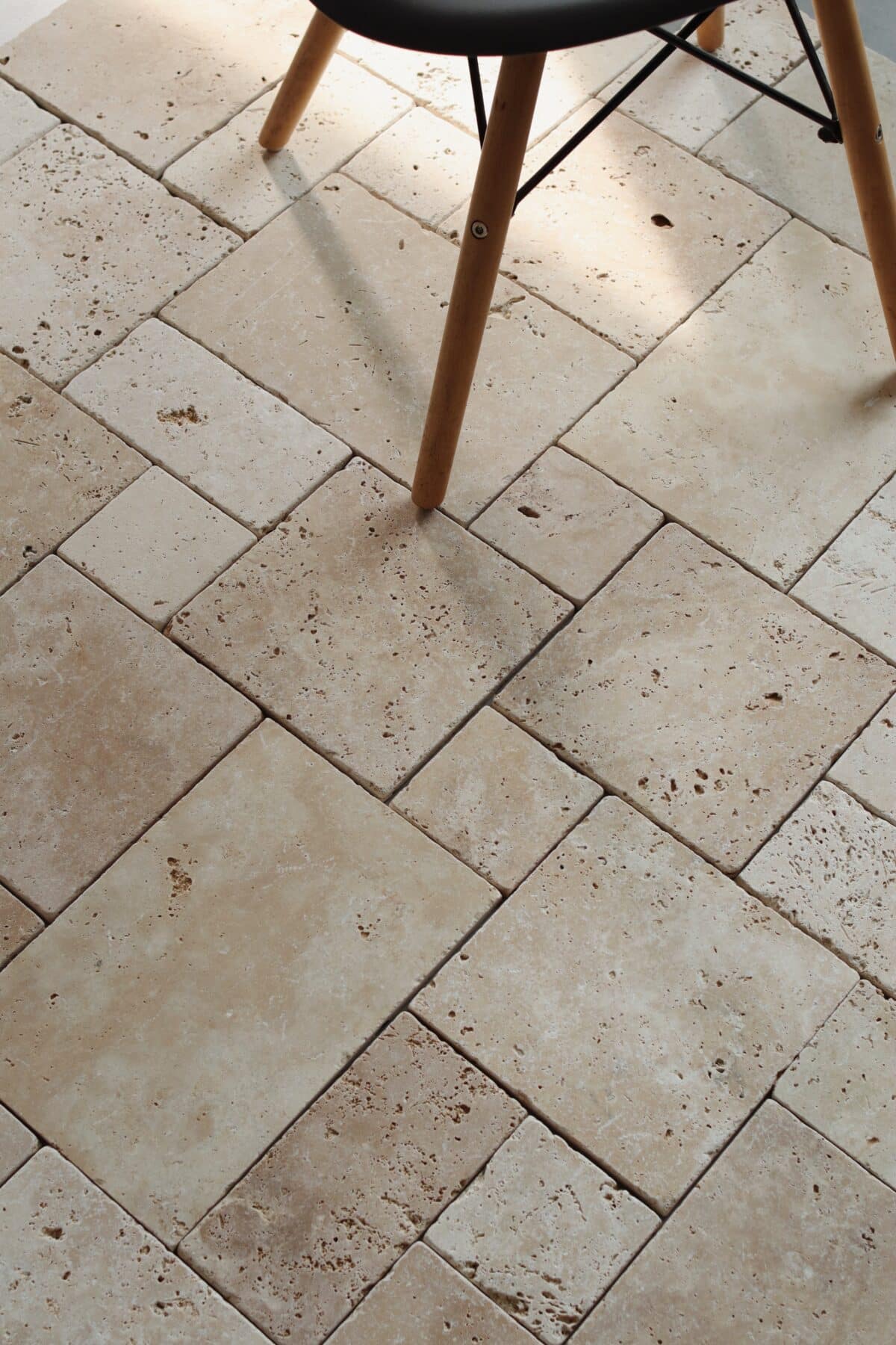 Podłoga z trawertynu IVORY MIX MINI w układzie rzymskim, w ciepłych odcieniach beżu, z widocznymi naturalnymi porami i delikatnym szlifowanym wykończeniem. Na pierwszym planie drewniane nogi czarnego krzesła na metalowej konstrukcji.