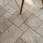 Podłoga z trawertynu IVORY MIX MINI w układzie rzymskim, w ciepłych odcieniach beżu, z widocznymi naturalnymi porami i delikatnym szlifowanym wykończeniem. Na pierwszym planie drewniane nogi czarnego krzesła na metalowej konstrukcji.