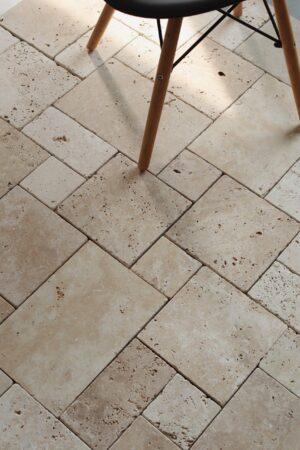 Podłoga z trawertynu IVORY MIX MINI w układzie rzymskim, w ciepłych odcieniach beżu, z widocznymi naturalnymi porami i delikatnym szlifowanym wykończeniem. Na pierwszym planie drewniane nogi czarnego krzesła na metalowej konstrukcji.