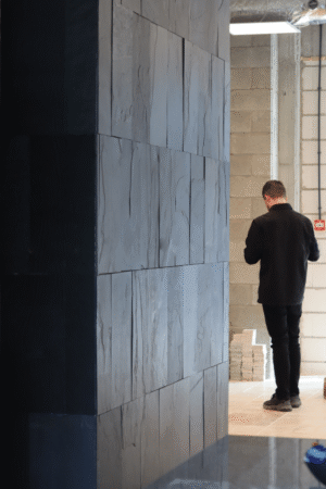 Wnętrze z czarną ścianą z łupka BLACK SLATE 30x60 cm – duży format płytek, łupana struktura i męska sylwetka w tle.