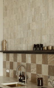 Jasne cegiełki z trawertynu Ivory 15x7,5 cm ułożone na ścianie nad kuchennym blatem, z ciemną półką i kontrastowymi akcentami.