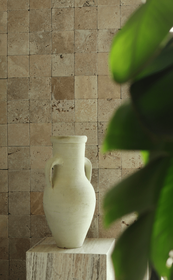 Ściana wyłożona płytkami trawertynowymi Ivory w formacie 10x10 cm, z dekoracyjnym dzbanem na postumencie z kamienia i liśćmi rośliny na pierwszym planie.