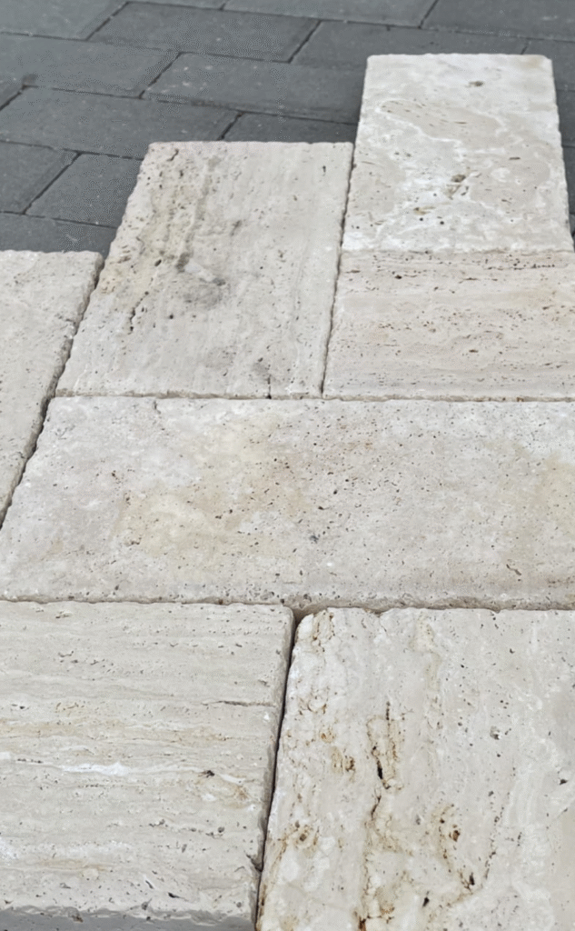 Perspektywiczny widok na jasne płytki z trawertynu Classic Light ułożone obok ciemnej kostki brukowej – naturalna struktura kamienia z porowatą, szczotkowaną powierzchnią.