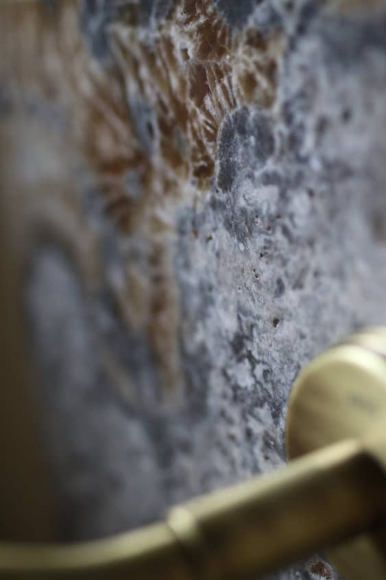 Makrozdjęcie ściany z kamienia Ash Mix z wyraźnym motywem skamieniałości i fragmentem złotej baterii – kontrast naturalnych tekstur i szlachetnego metalu.