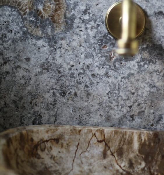 Zbliżenie na ścianę z kamienia naturalnego Ash Mix z widoczną skamieniałością w tle, złotym uchwytem i brązowo-beżową umywalką z kamienia.
