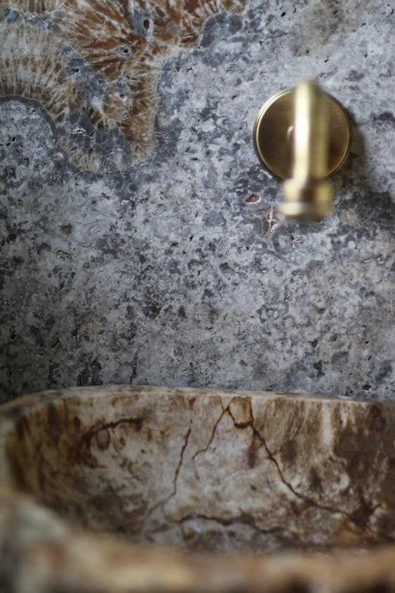 Zbliżenie na ścianę z kamienia naturalnego Ash Mix z widoczną skamieniałością w tle, złotym uchwytem i brązowo-beżową umywalką z kamienia.