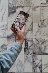 Ręka trzymająca telefon nad płytkami z marmuru Lilac uchwyconymi z bliska. Dynamiczne użylenie w odcieniach fioletu i czerni na tle bieli podkreśla unikatowość naturalnego kamienia.
